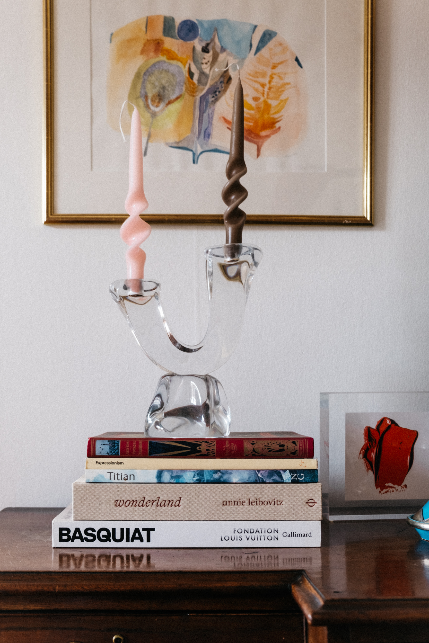 A vintage organic crystal chandelier with pink and brown twisted candles atop a pile of art books in front of a framed painting next to a piece by the artist CJ Henry
