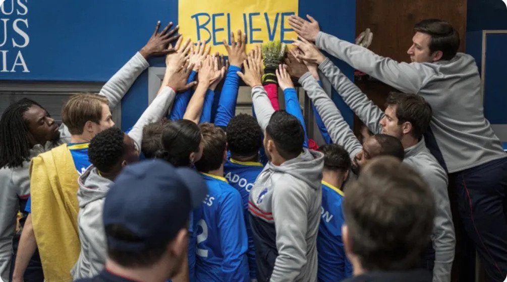 A picture of an episode of Ted Lasso where all the players are touching the handwritten poster reading "Believe"
